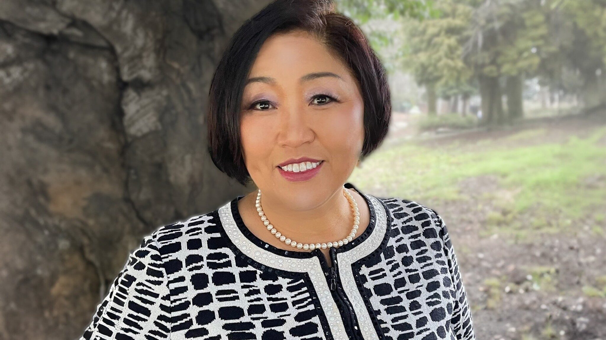 Cheryl-Lee_16x9-min Woman in a patterned jacket and uncommon thinkers pearl necklace smiling outdoors.