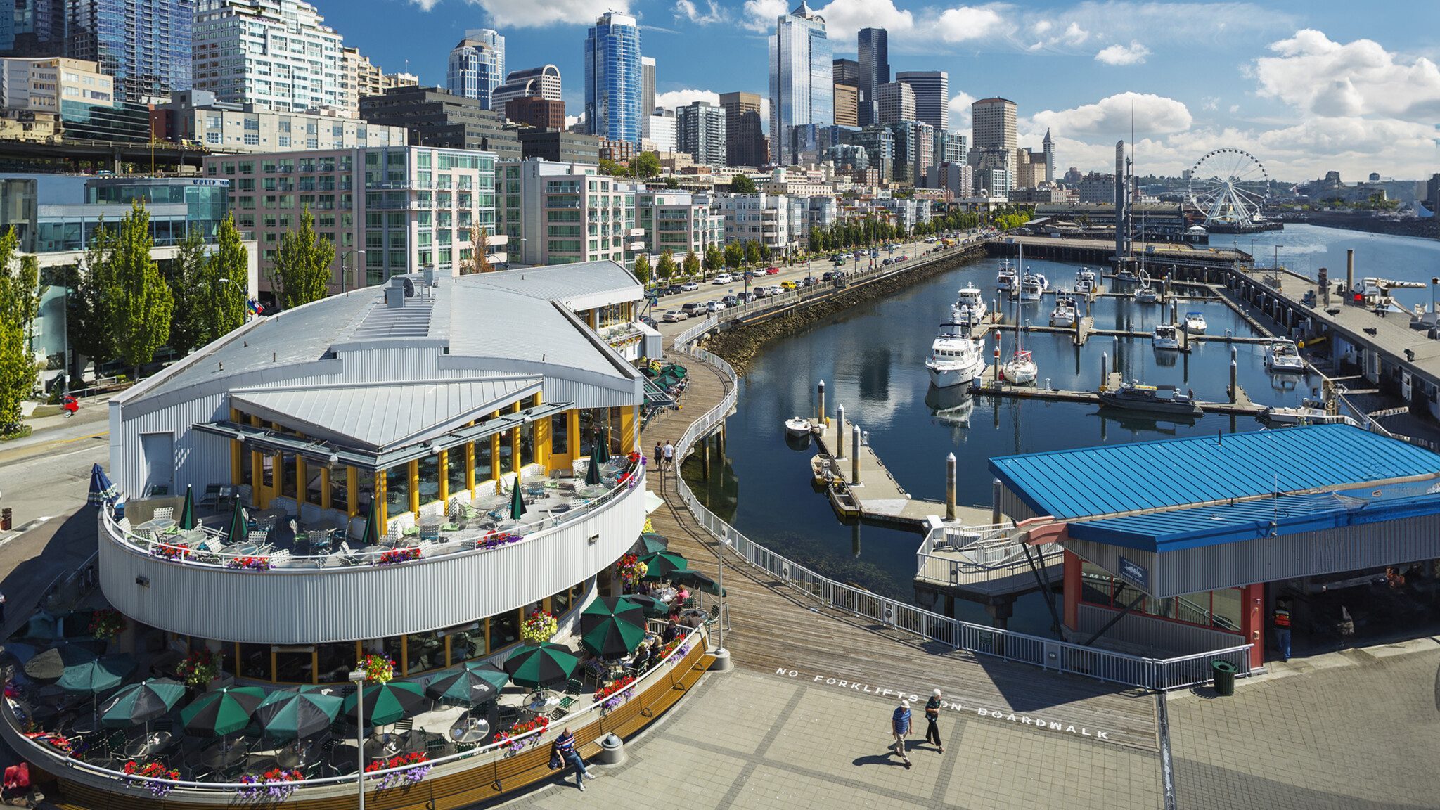 SeattleWaterfrontPark_16x9 A bustling waterfront with a restaurant terrace overlooking a marina against a backdrop of Seattle's modern city skyline, enhancing tourism.